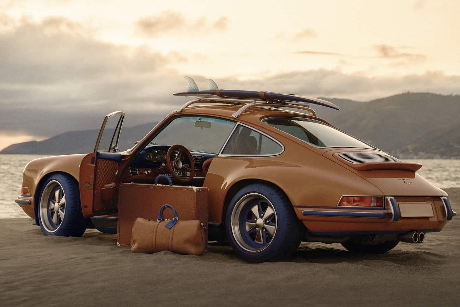 Side exterior profile showing carbon fibre bodywork presented in Saffron, with roof rack and surfboard fitted. Shown with Louis Vuitton luggage collection.